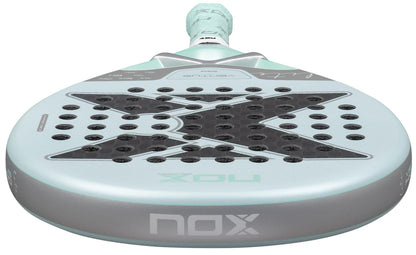 A close-up of the NOX Luxury Ventus 12K Hybrid Lite 2026 Padel Racket shows its light blue face with a bold black "X," multiple holes, premium 12K carbon fiber construction, and the handle visible at the top.