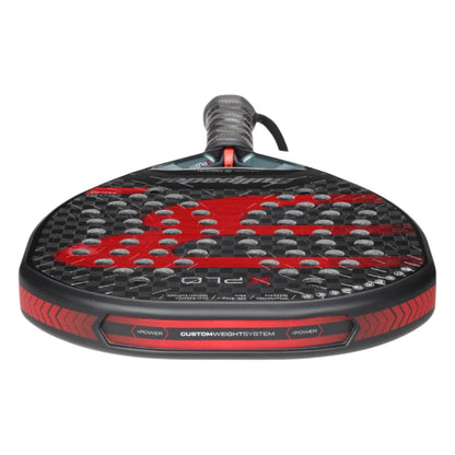 A close-up of the Bullpadel Xplo 2025 Padel Racket highlights its black and red design, perforated face, textured grip handle, and Bullpadel branding with detailed text on the frame.