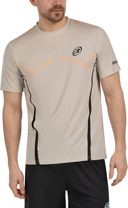 A man wears a Bullpadel Cabore Men's Padel T-Shirt in Peanut with Quickerdry tech and black graphics, paired with black shorts, standing with arms relaxed at his sides.