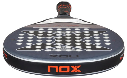 A close-up of the NOX AT10 Pro Cup Comfort By Agustin Tapia 2025 padel racket shows its round head with white holes, black and gray geometric patterns, the orange NOX logo on the edge, and a visible Smartstrap® for safety.
