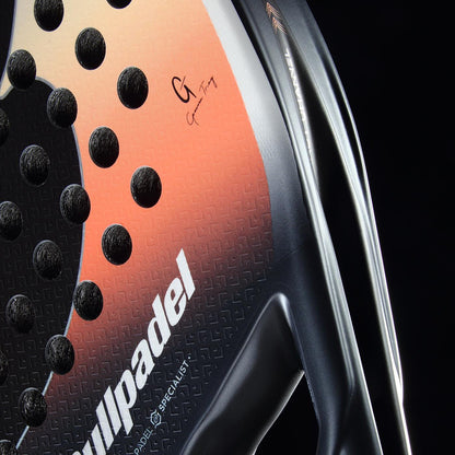 Close-up of the Bullpadel Elite W 2026 Padel Racket by Bullpadel, a lightweight racket featuring a black and orange gradient, circular holes, textured surface, and a visible signature on the upper right side.