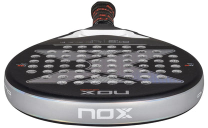 A close-up of the black NOX AT10 Pro Cup HARD By Agustin Tapia 2025 padel racket, featuring a silver edge, X-pattern holes, and a red-black grip handle with Smartstrap® system for safety.
