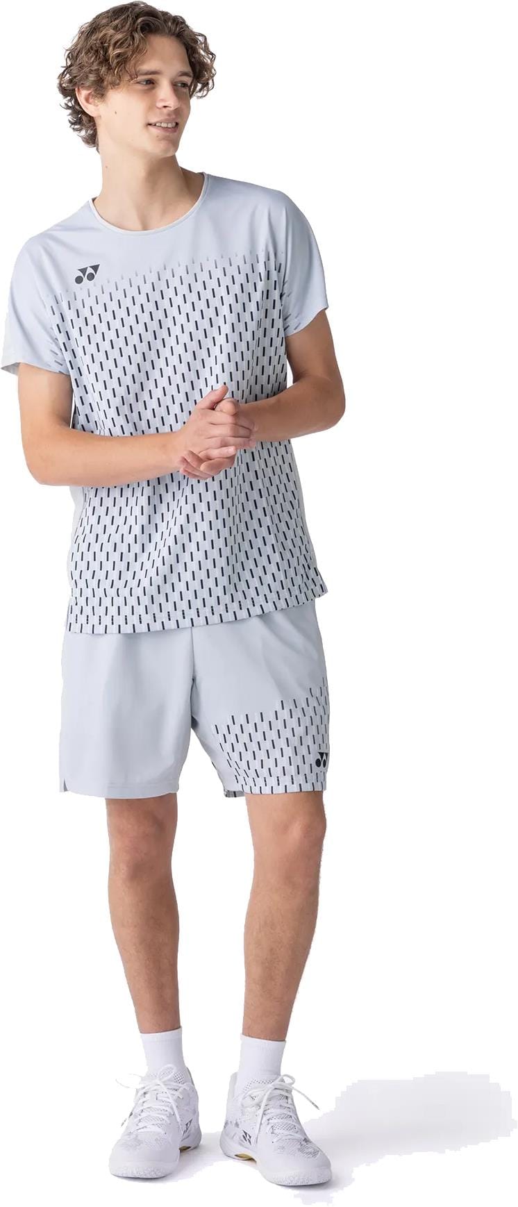 A young man stands wearing Yonex 15241EX Knit Mens Padel Shorts in Silver Grey, paired with a matching shirt and white sneakers. He looks to the side, hands clasped together in a relaxed pose.