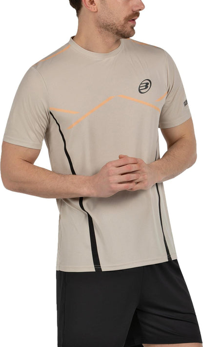A man wearing a light beige Bullpadel Cabore Mens Padel T-Shirt - Peanut with orange accents and a chest logo, paired with black shorts, stands slightly turned to the side with his hands clasped.