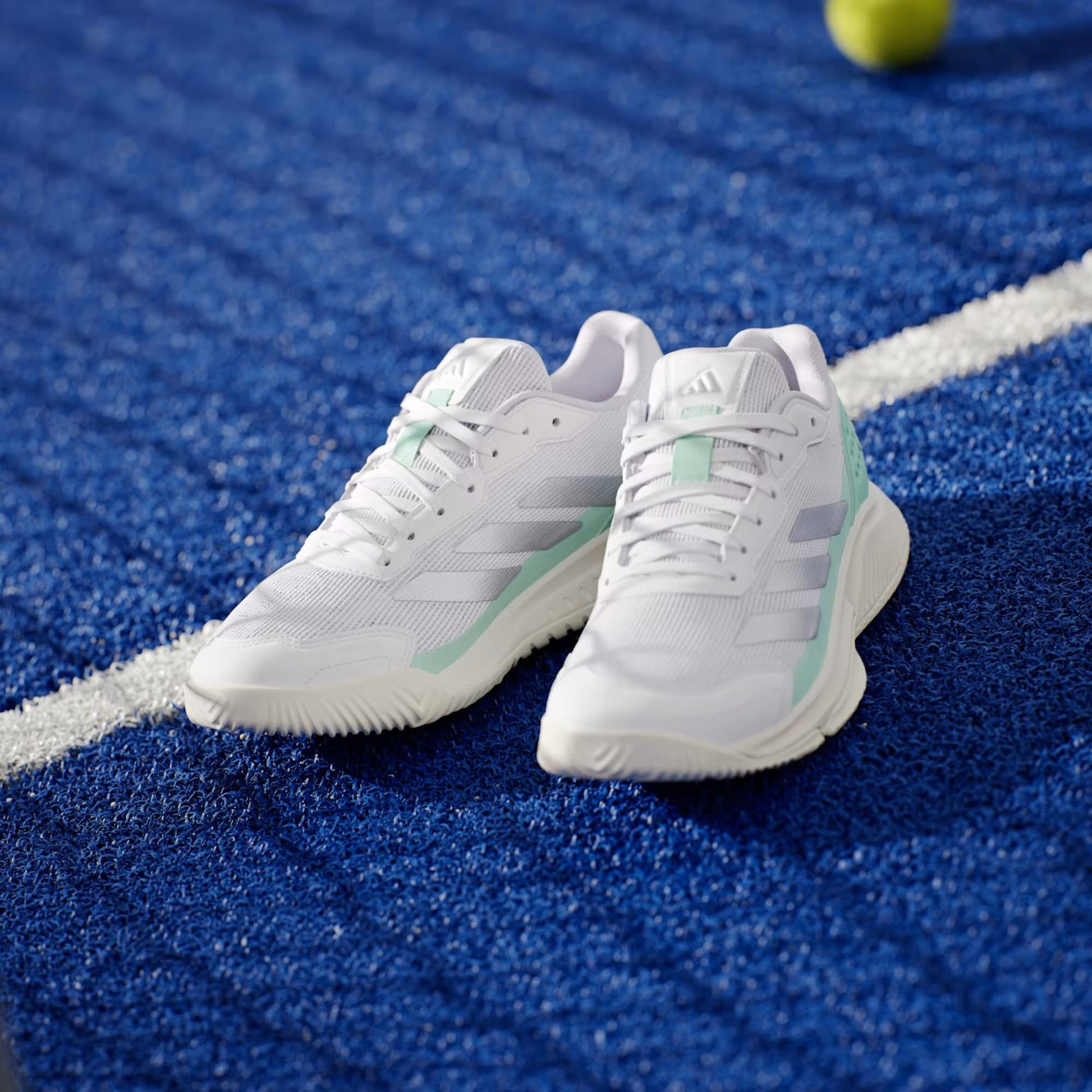 The adidas Courtquick Womens Padel Shoes in Cloud White, Silver Metallic, and Clear Mint rest on a blue court with a tennis ball nearby—ideal stylish performance footwear for padel players.