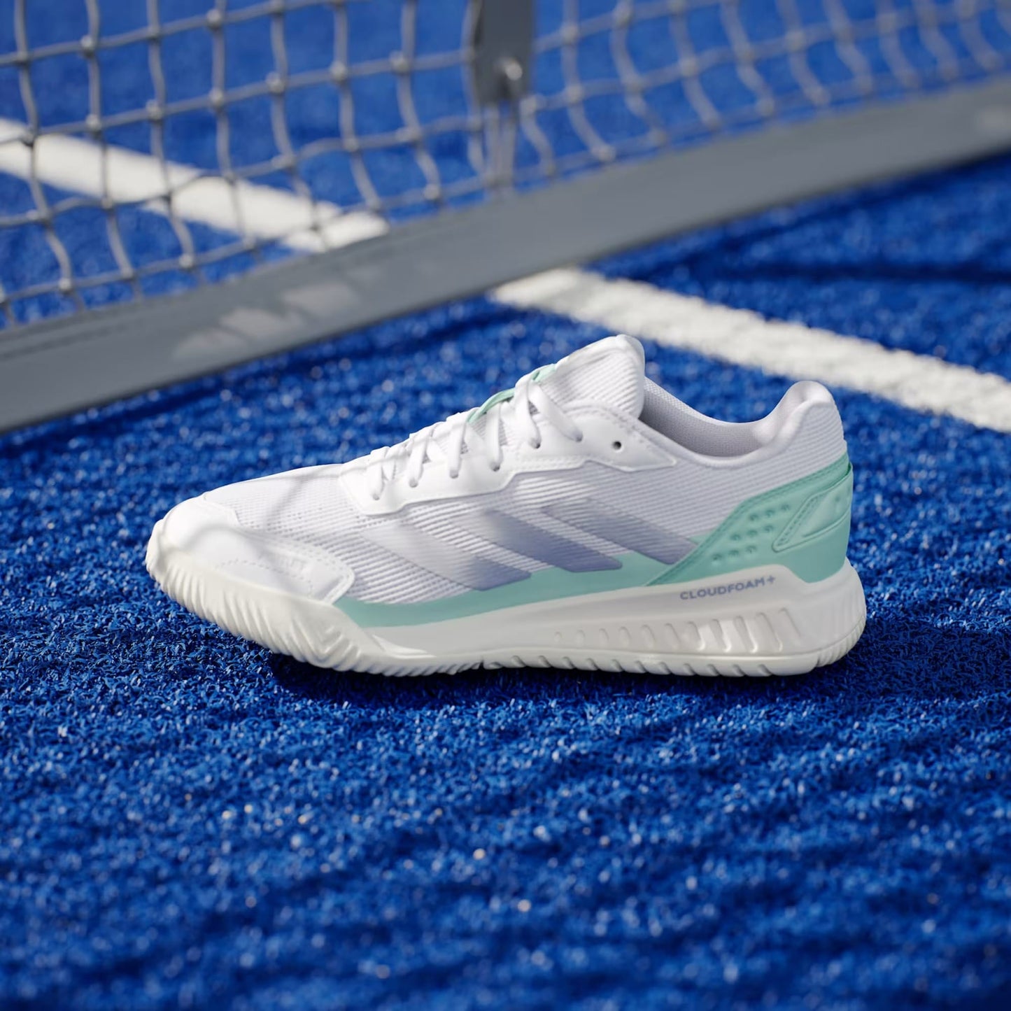 The adidas Courtquick Womens Padel Shoes in Cloud White, Silver Metallic, and Clear Mint rest on a blue tennis court near a white line, with the net visible in the background. Cloudfoam+ branding is featured.